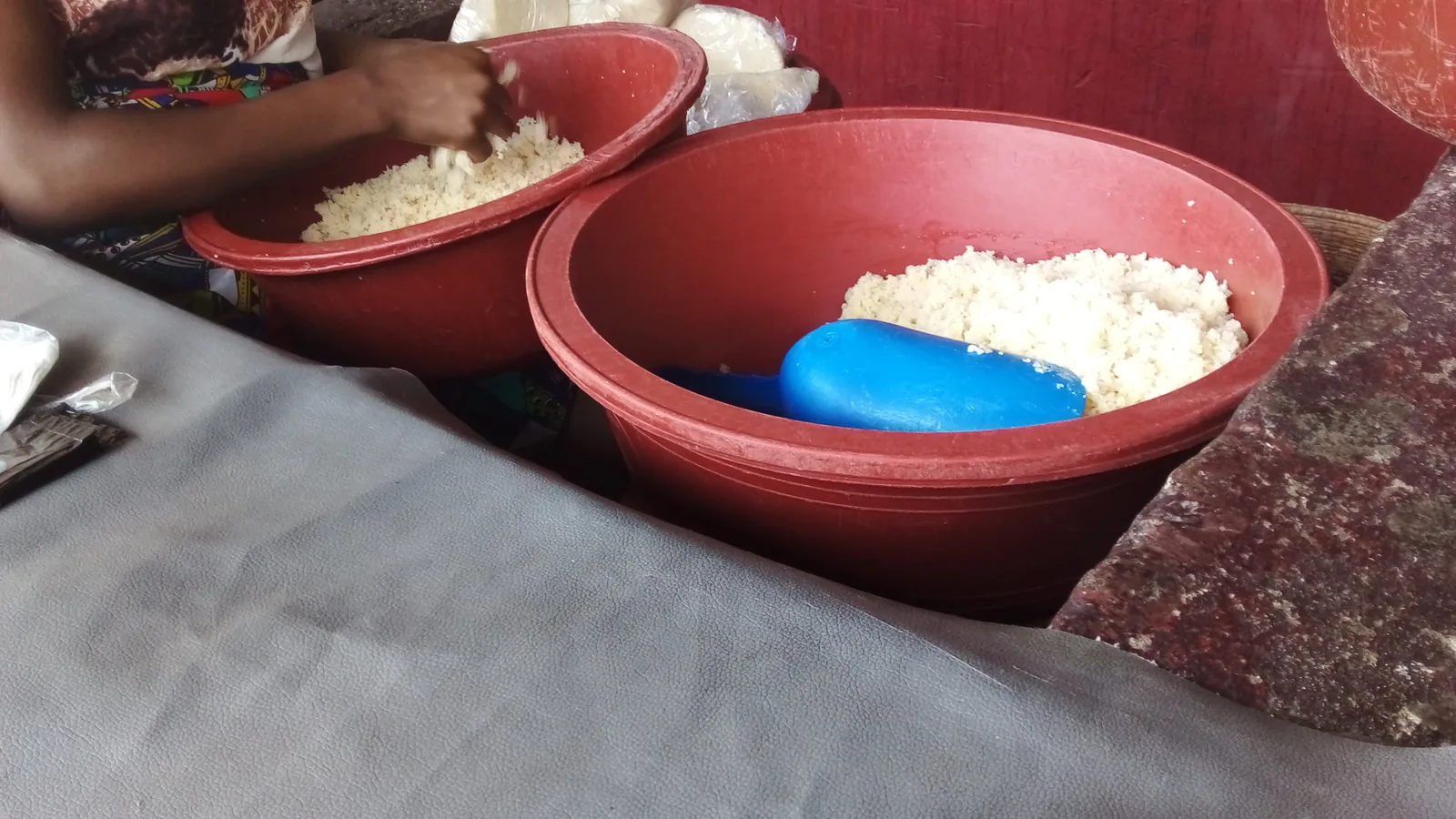 Two red plastic basins of loose attiéké on a market stall with a blue plastic scoop and a vendor's hand