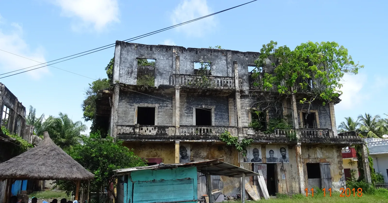 A roofless colonial house in the Quartier France at Grand-Bassam, its veranda balustrades intact and trees growing out of the ruin, with pasted portrait artworks on the ground floor
