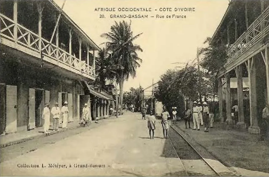 A colonial-period postcard captioned AFRIQUE OCCIDENTALE — COTE D'IVOIRE, GRAND-BASSAM, RUE DE FRANCE, showing verandaed buildings on both sides of a street with a palm, tram rails and figures