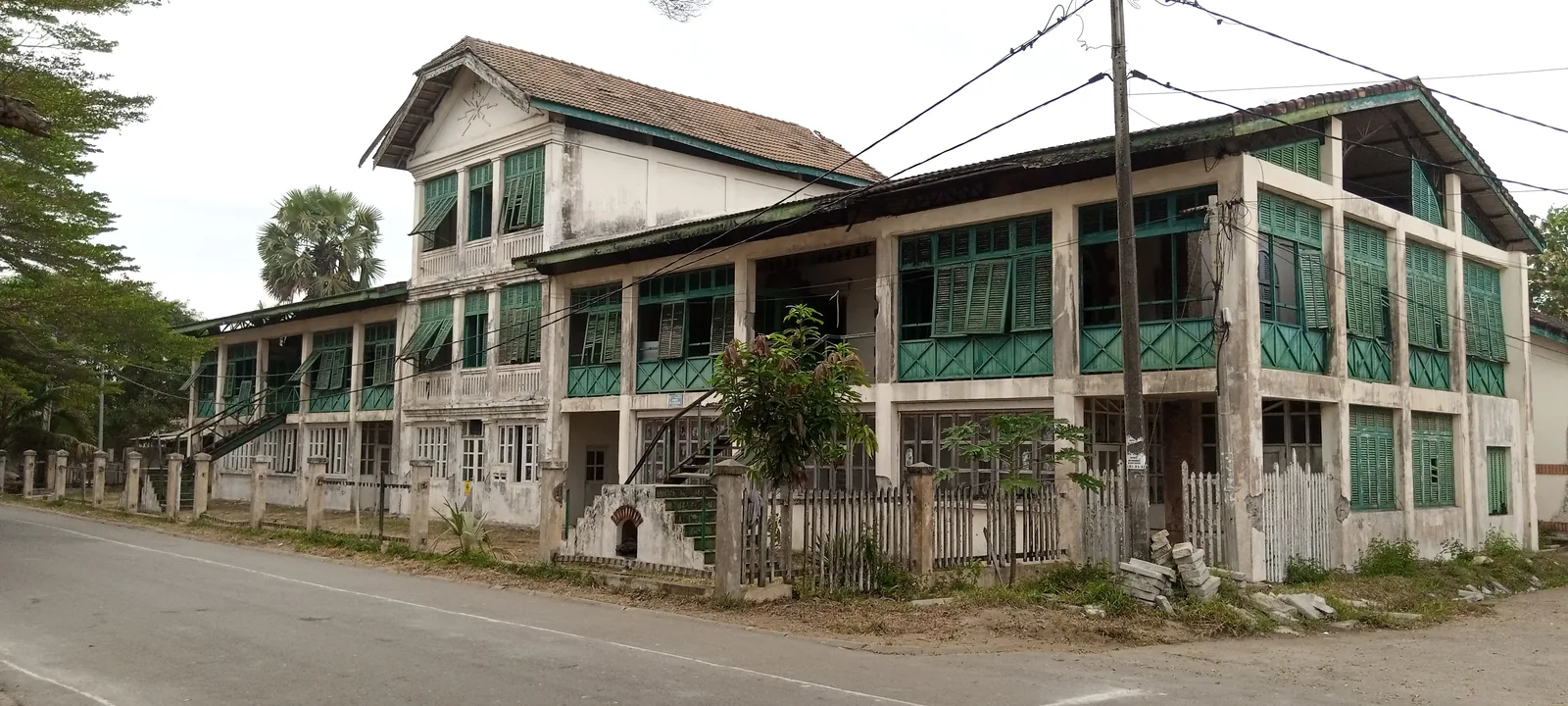 A long two-storey colonial house with green louvred shutters and verandas on an empty road at Grand-Bassam