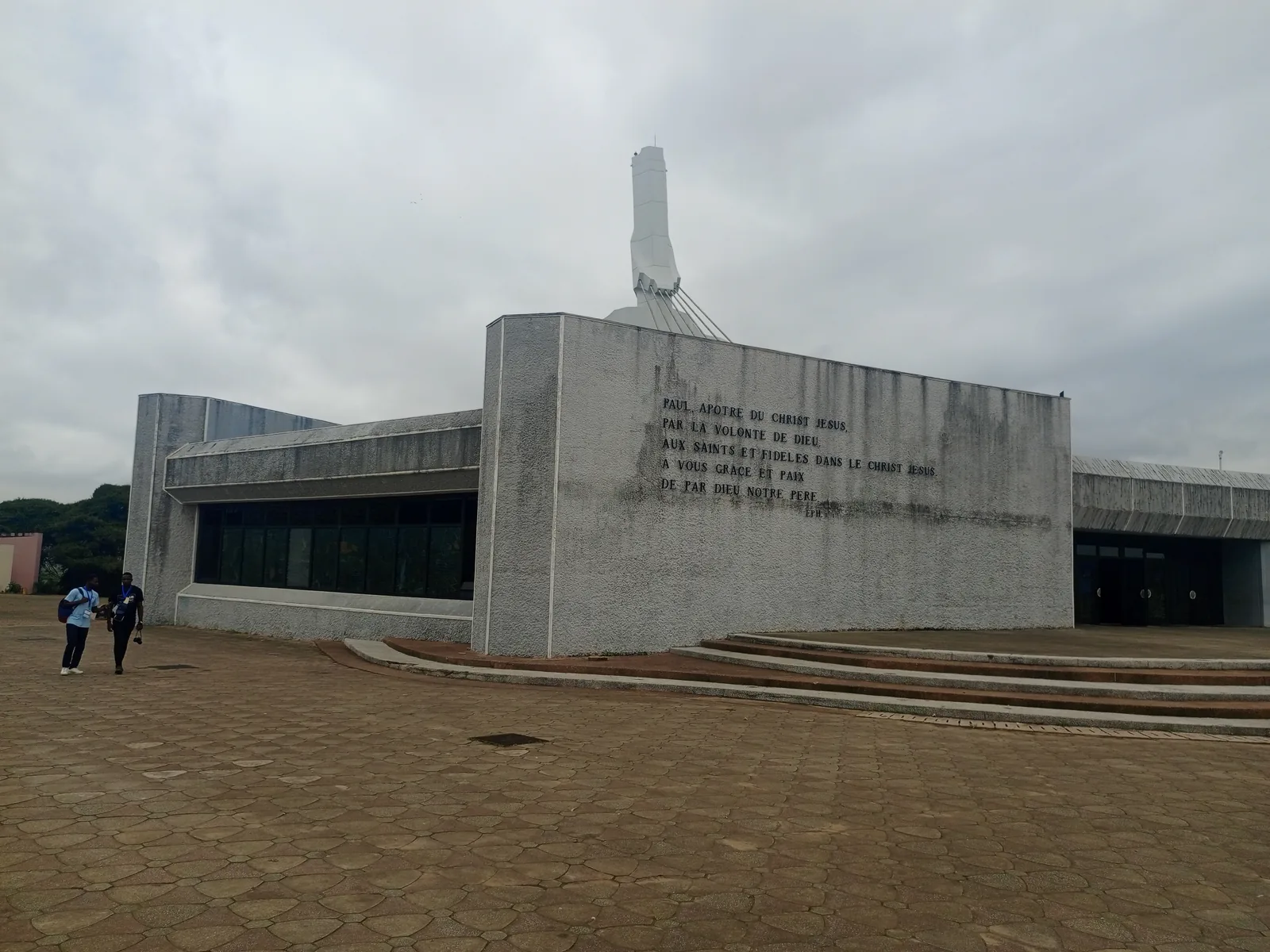 The blank inscribed side wall of St Paul's Cathedral, Abidjan, with the tip of the white pylon showing above it and an empty paved forecourt in front