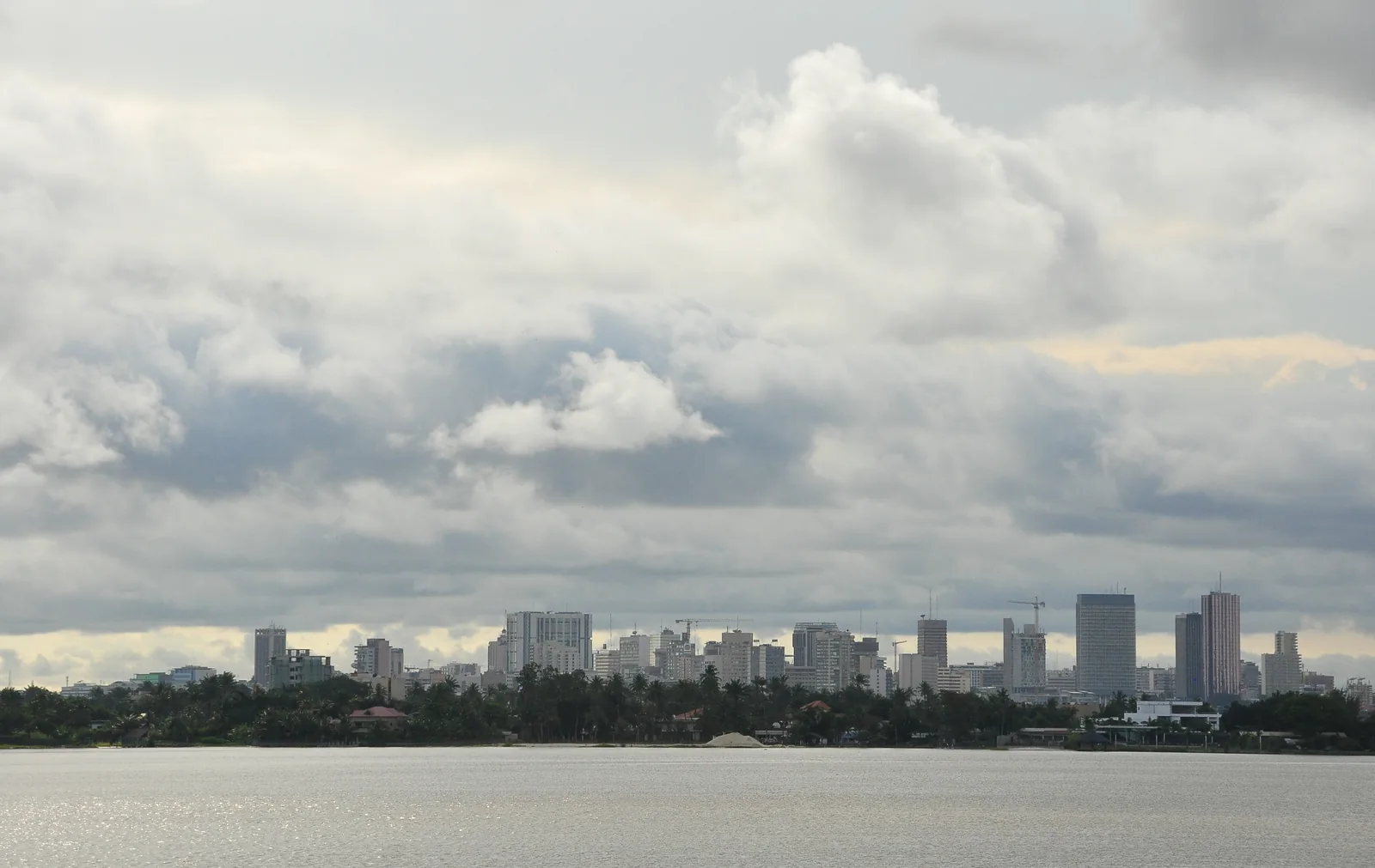 The Plateau skyline of Abidjan seen across the lagoon from the water, a line of towers and construction cranes above a fringe of palms under heavy cloud
