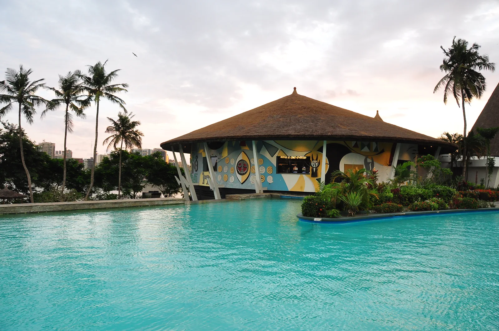The swimming pool and a thatched, mask-muralled bar pavilion in the grounds of the Hôtel Ivoire at dusk, with the Plateau skyline faint in the distance