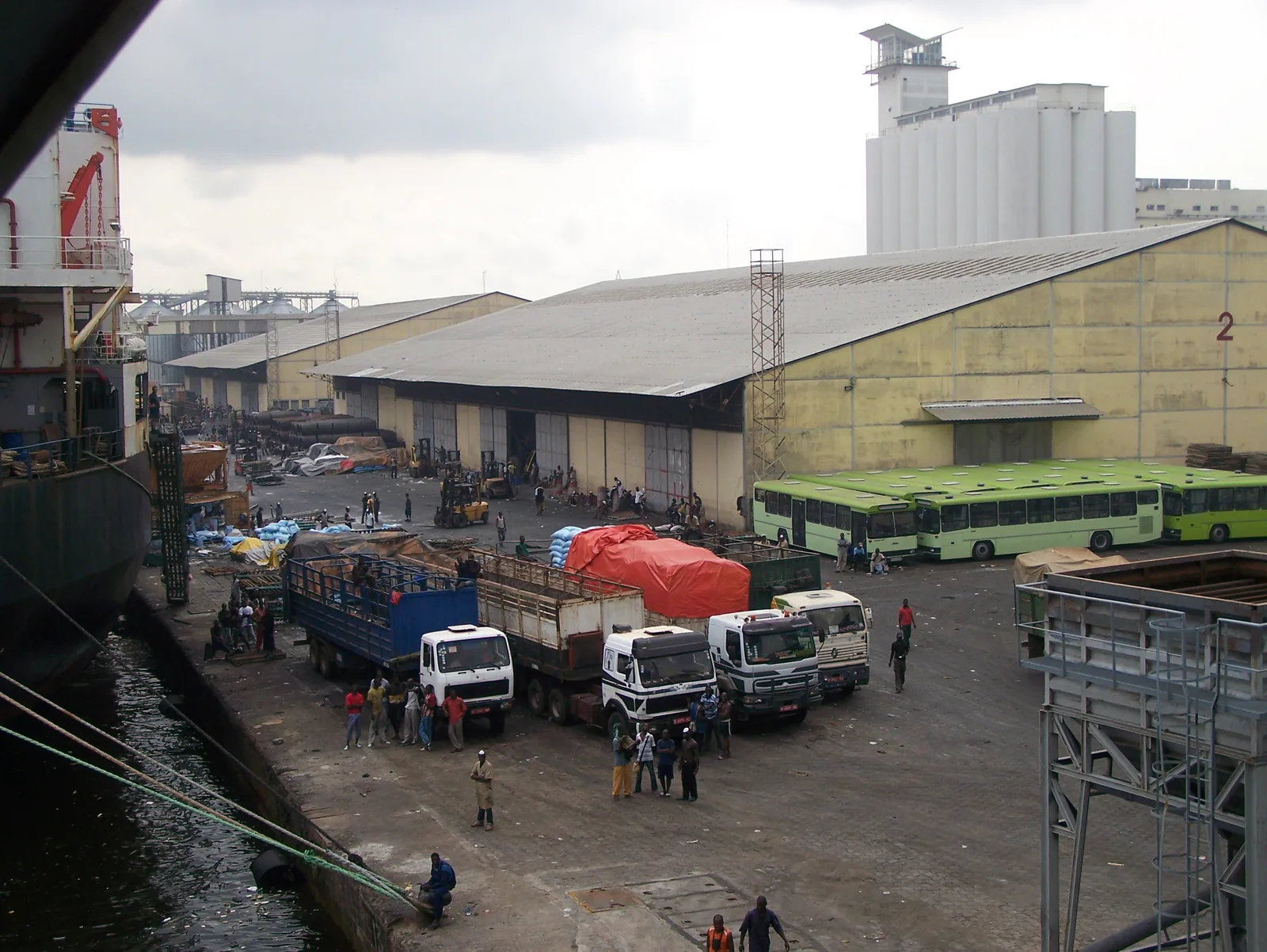 A working cargo quay at the Port of Abidjan seen from a ship's deck, with a warehouse shed, grain silos, trucks and dockers handling bagged cargo