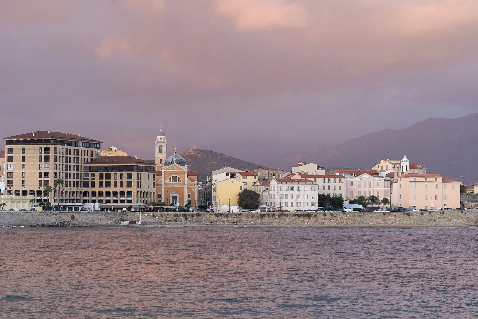 Ajaccio waterfront at sunset with cathedral dome, pastel buildings, and Corsican mountains in pink-purple light