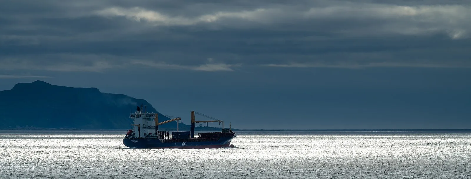 Cargo ship silhouetted in silver Norwegian Sea with dramatic clouds and mountain headland