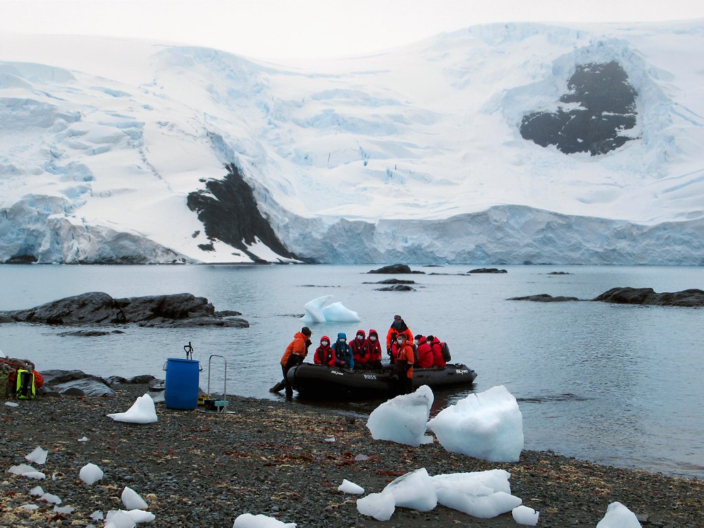 Zodiac inflatable boat with passengers in red parkas landing on Antarctic shore with glaciers behind