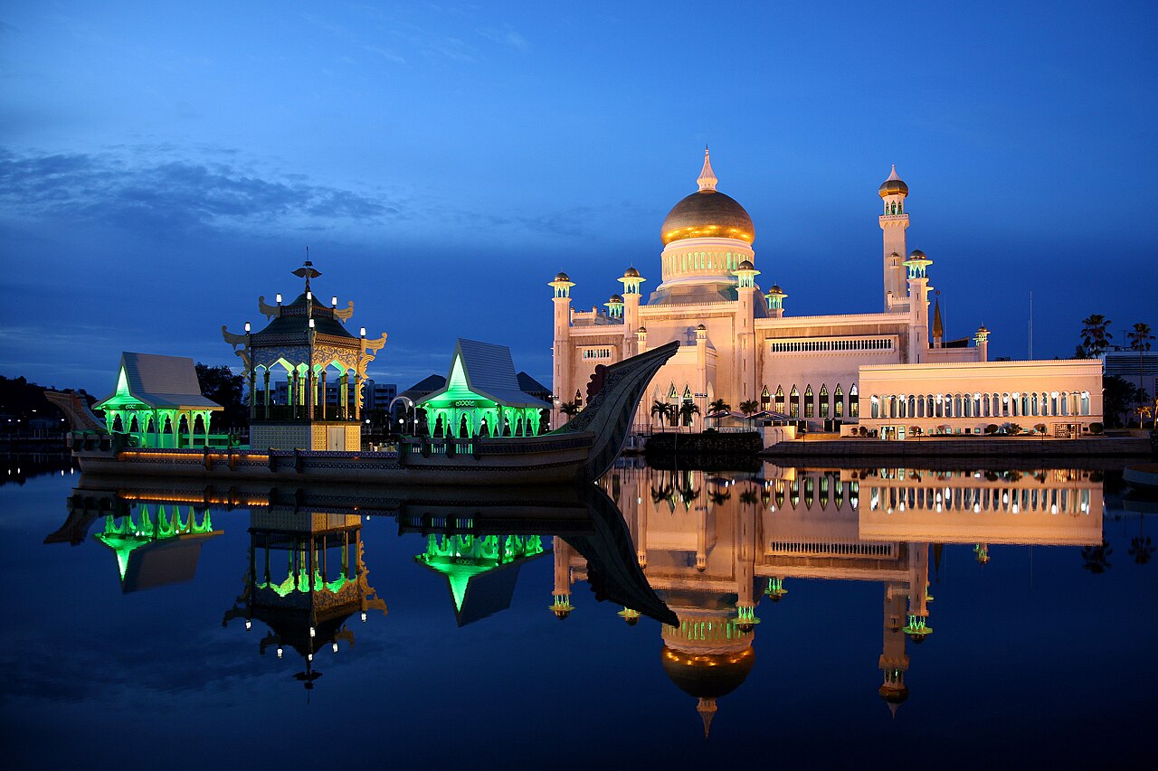 Omar Ali Saifuddien Mosque illuminated at blue hour with golden dome and green-lit barge reflected in lagoon