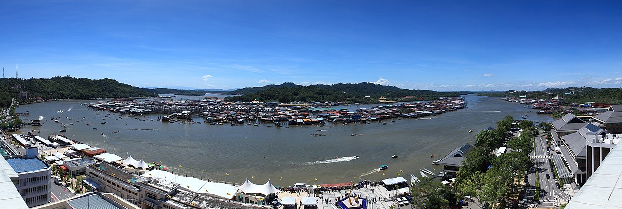 Wide panorama of Kampong Ayer water village on the Brunei River, with city waterfront and forested hills beyond