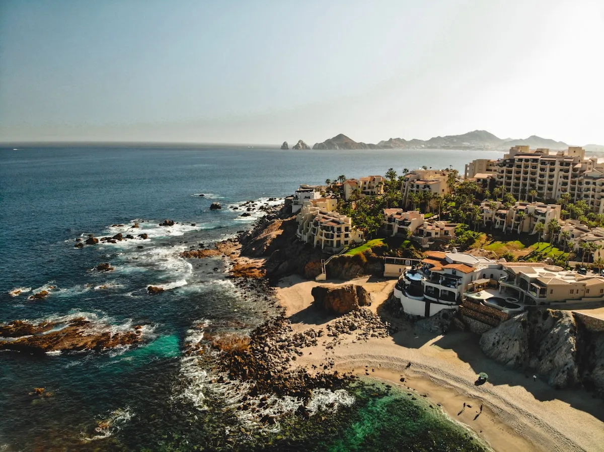 Aerial view of the Cabo San Lucas coastline showing resort buildings along rocky cliffs, small beach cove, and Land's End rock formations in the hazy distance