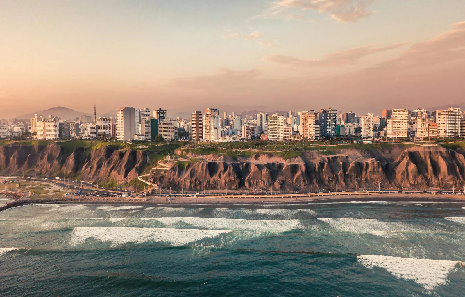 Aerial sunset view of the Miraflores skyline of apartment towers standing along the top of the Costa Verde cliffs in Lima, with the coastal highway at the cliff base and Pacific surf rolling onto the beach in the foreground