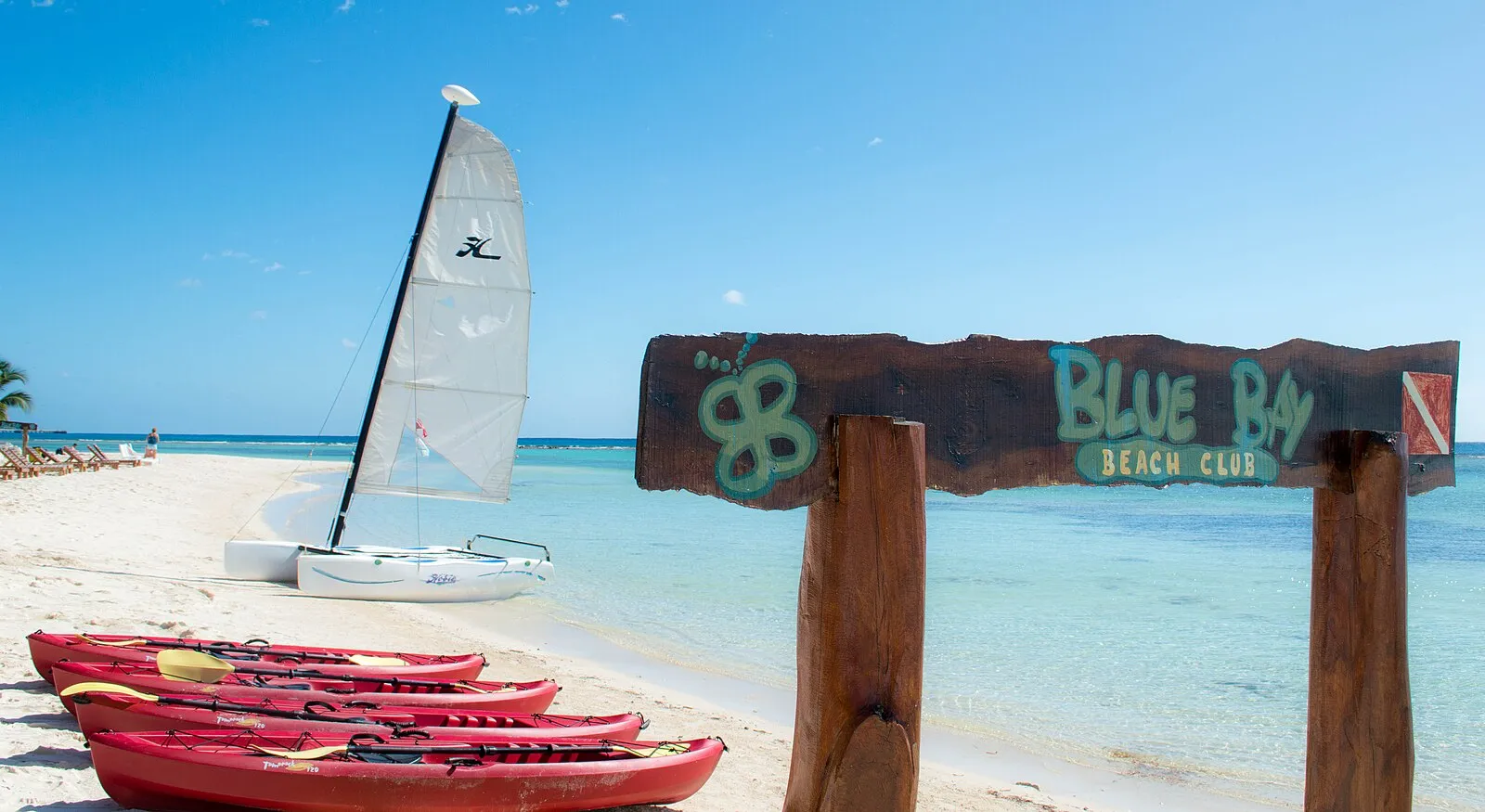 Blue Bay Beach Club sign with kayaks and Hobie Cat on white sand