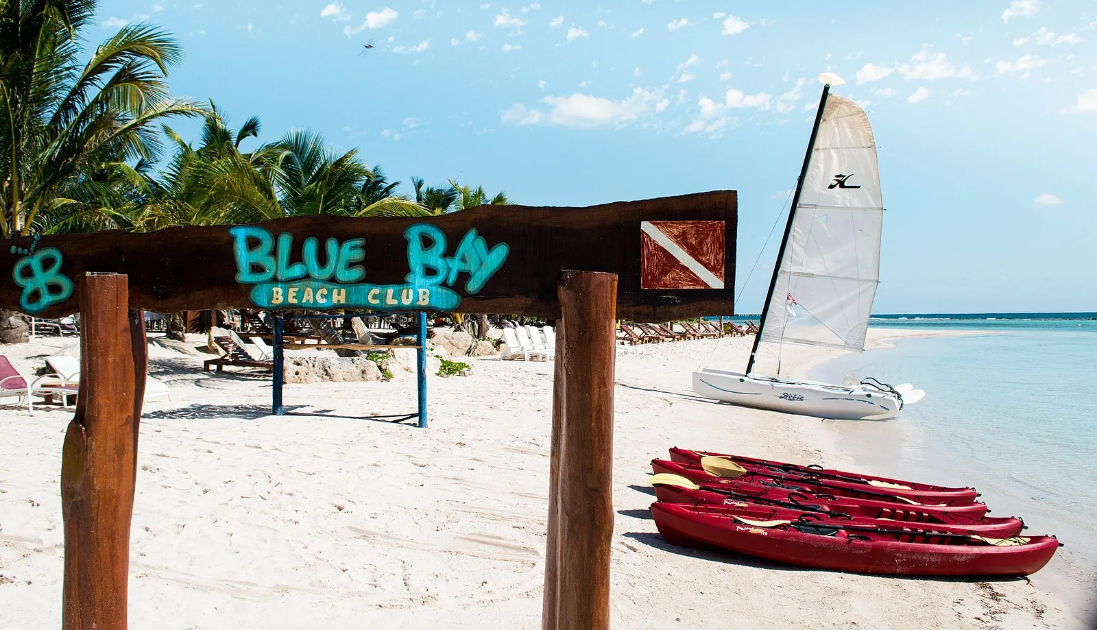 Blue Bay Beach Club sign on white sand beach with kayaks and Hobie Cat sailboat