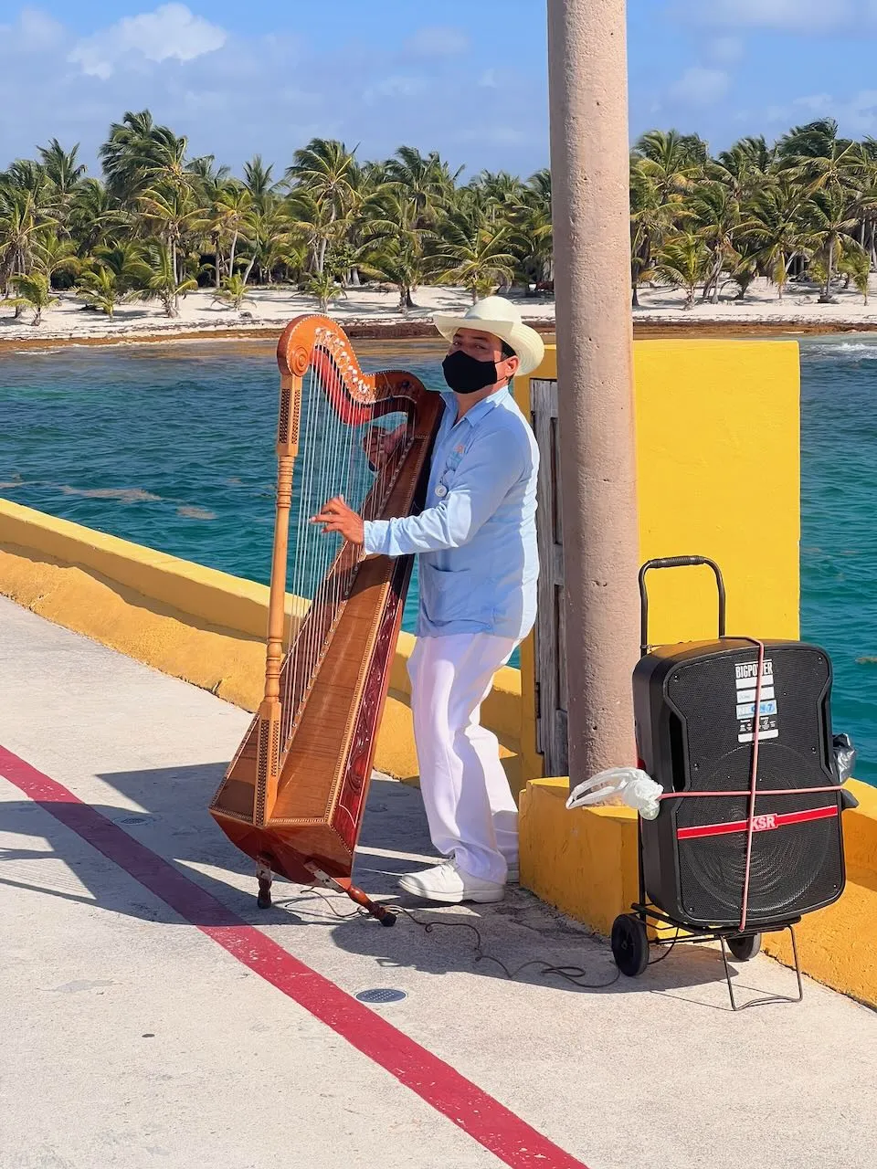 Local harp musician in white traditional clothing playing on the Costa Maya pier with palm trees and turquoise water behind him