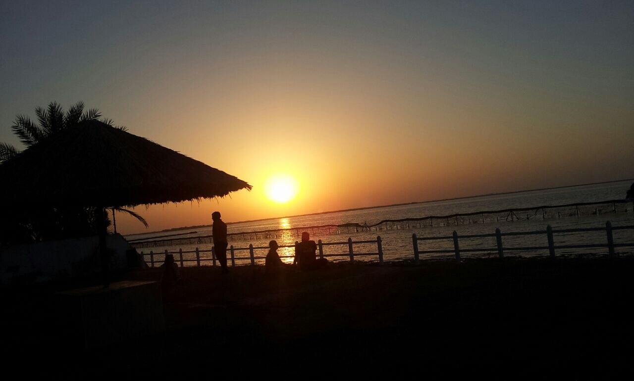 Beachside sunset with thatched palm umbrella and people watching the sun set over the Arabian Gulf