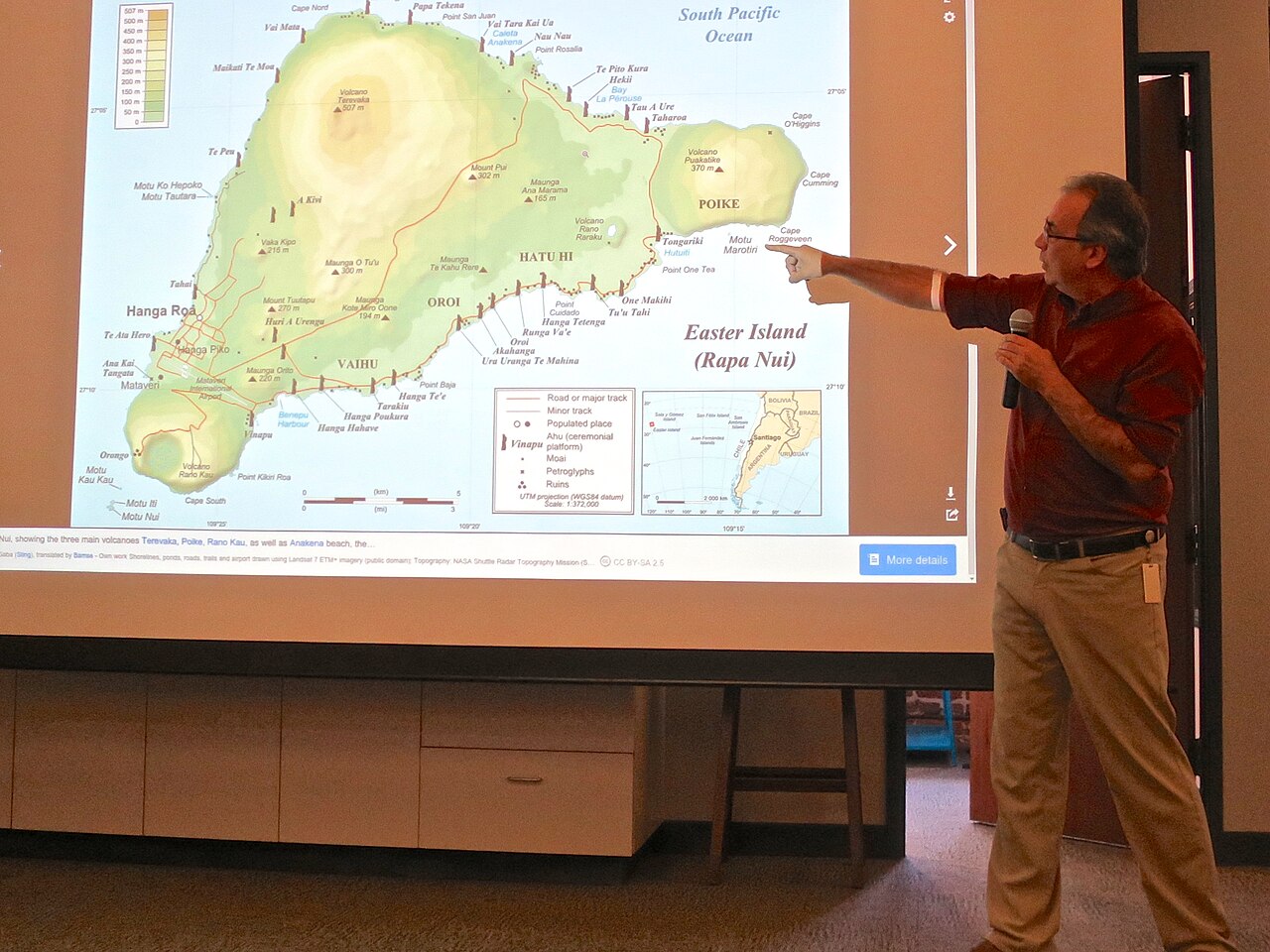 Presenter pointing at a detailed topographic map of Easter Island (Rapa Nui) showing volcanoes, moai sites, and roads