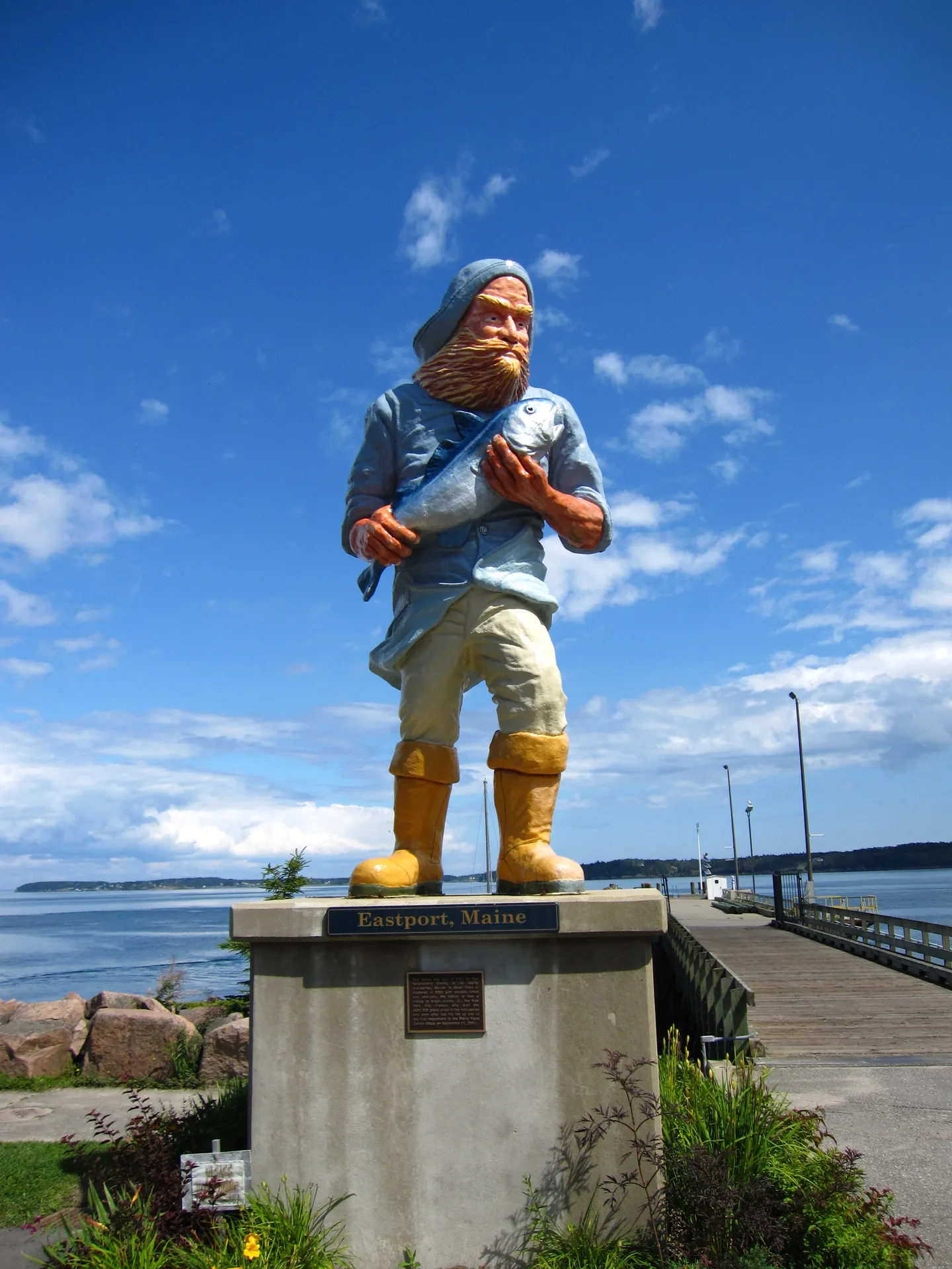A tall painted fisherman statue in oilskins and yellow boots, cradling a fish, standing on a pedestal reading 'Eastport, Maine' at the head of the breakwater pier, with Passamaquoddy Bay and a forested far shore behind under a blue sky