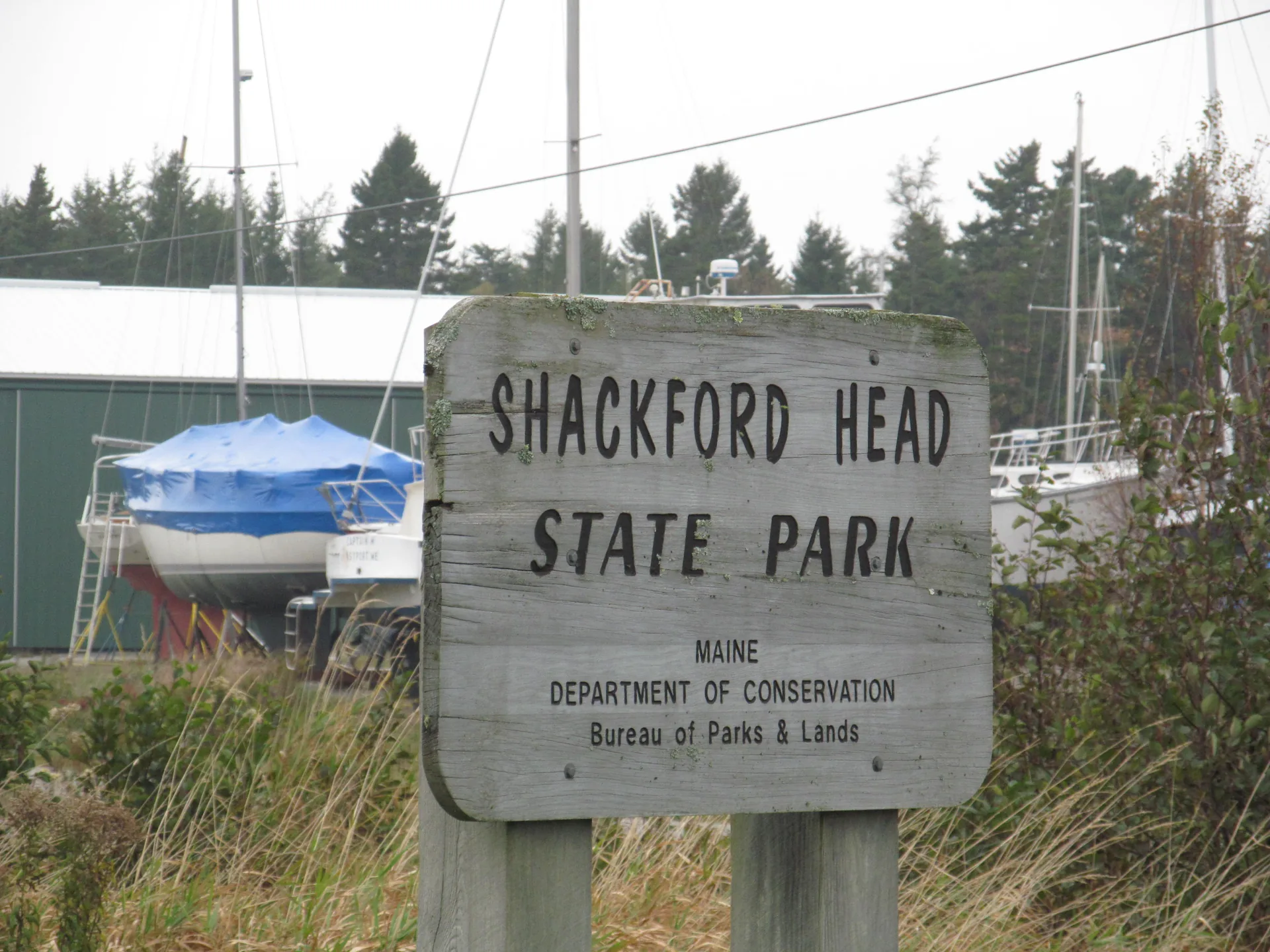 The weathered wooden entrance sign for Shackford Head State Park, Maine Department of Conservation, with sailboats stored on the hard behind it against a backdrop of spruce trees