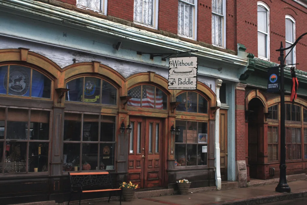Historic brick and painted storefronts along Water Street in downtown Eastport, with an antiques-and-art shop sign, an American flag in a shopfront window, a bench, and a cast-iron street lamp in the historic district