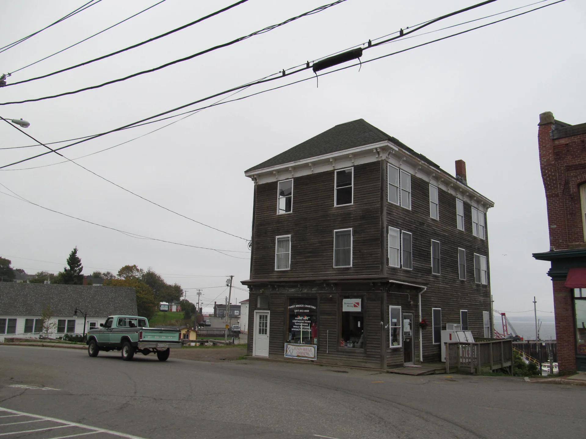 A weathered three-story wooden building near the Eastport waterfront housing a boat-charter and dive shop, with a vintage pickup truck passing and the bay visible beyond
