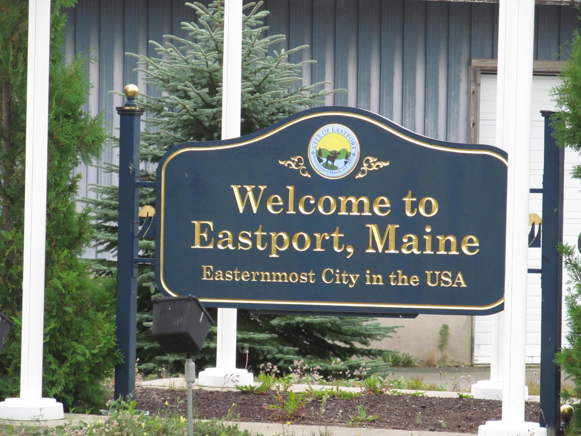 A dark blue city welcome sign lettered in gold reading 'Welcome to Eastport, Maine — Easternmost City in the USA,' topped by the city seal, set among young spruce trees