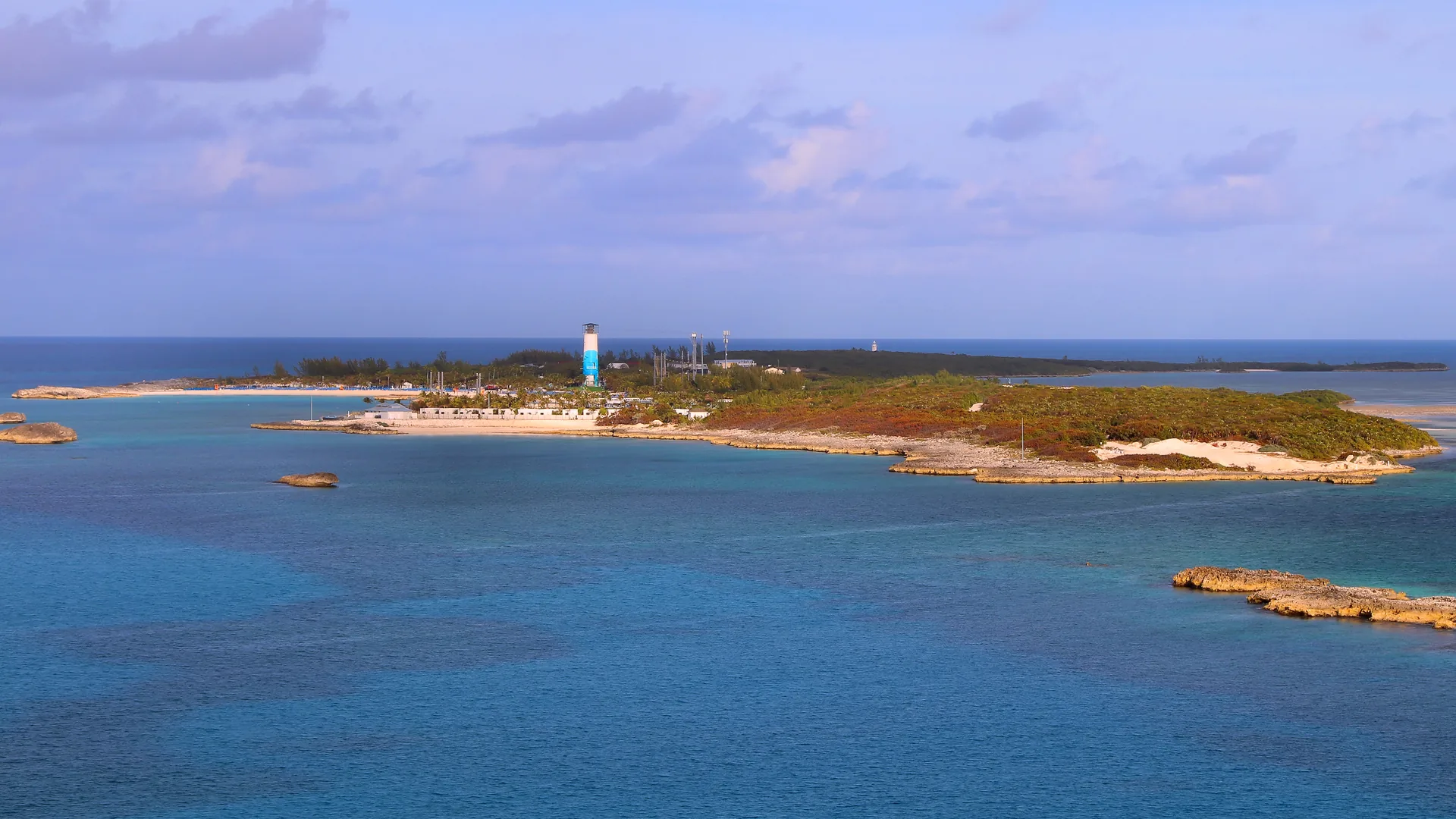 Great Stirrup Cay seen from an approaching ship: a low scrub-covered Bahamian island ringed by pale beach and turquoise water, with a tall tower over the developed area and the lighthouse on the far point