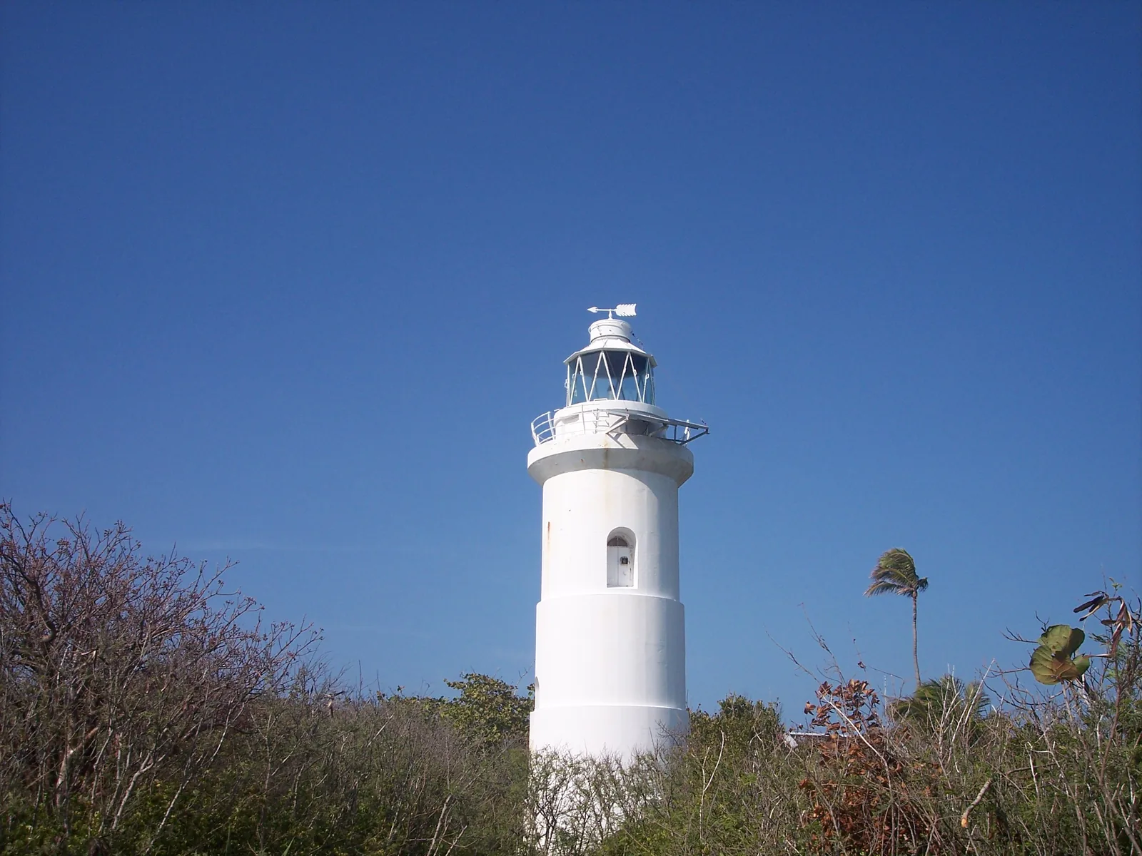 The white-painted Great Stirrup Cay lighthouse rising above low coastal scrub, its lantern room and gallery railing topped by an arrow weathervane under deep blue sky