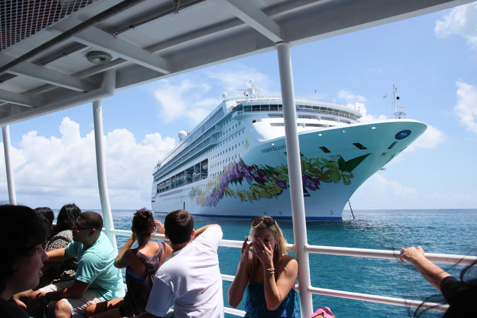 Passengers on a shaded tender deck watching the anchored Norwegian Sky across turquoise water