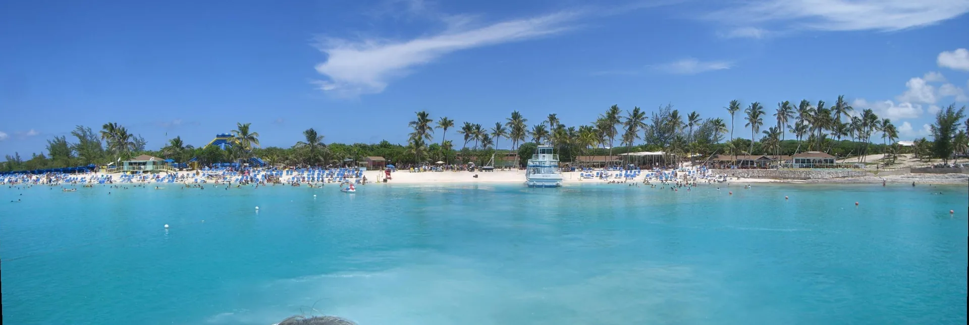 A wide panorama of the main beach at Great Stirrup Cay: a crescent of white sand lined with blue loungers and umbrellas, crowds swimming in shallow turquoise water, palms along the tree line, and a waterslide tower at left