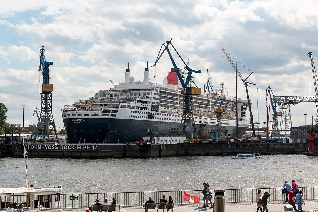 Queen Mary 2 in drydock at Blohm+Voss Dock Elbe 17 in Hamburg, with shipyard cranes and tourists walking along the Elbe promenade