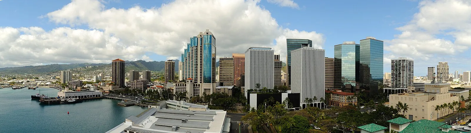 Panoramic view of downtown Honolulu skyline from the harbor with glass towers, palm trees, and the Ko'olau Mountains in the background
