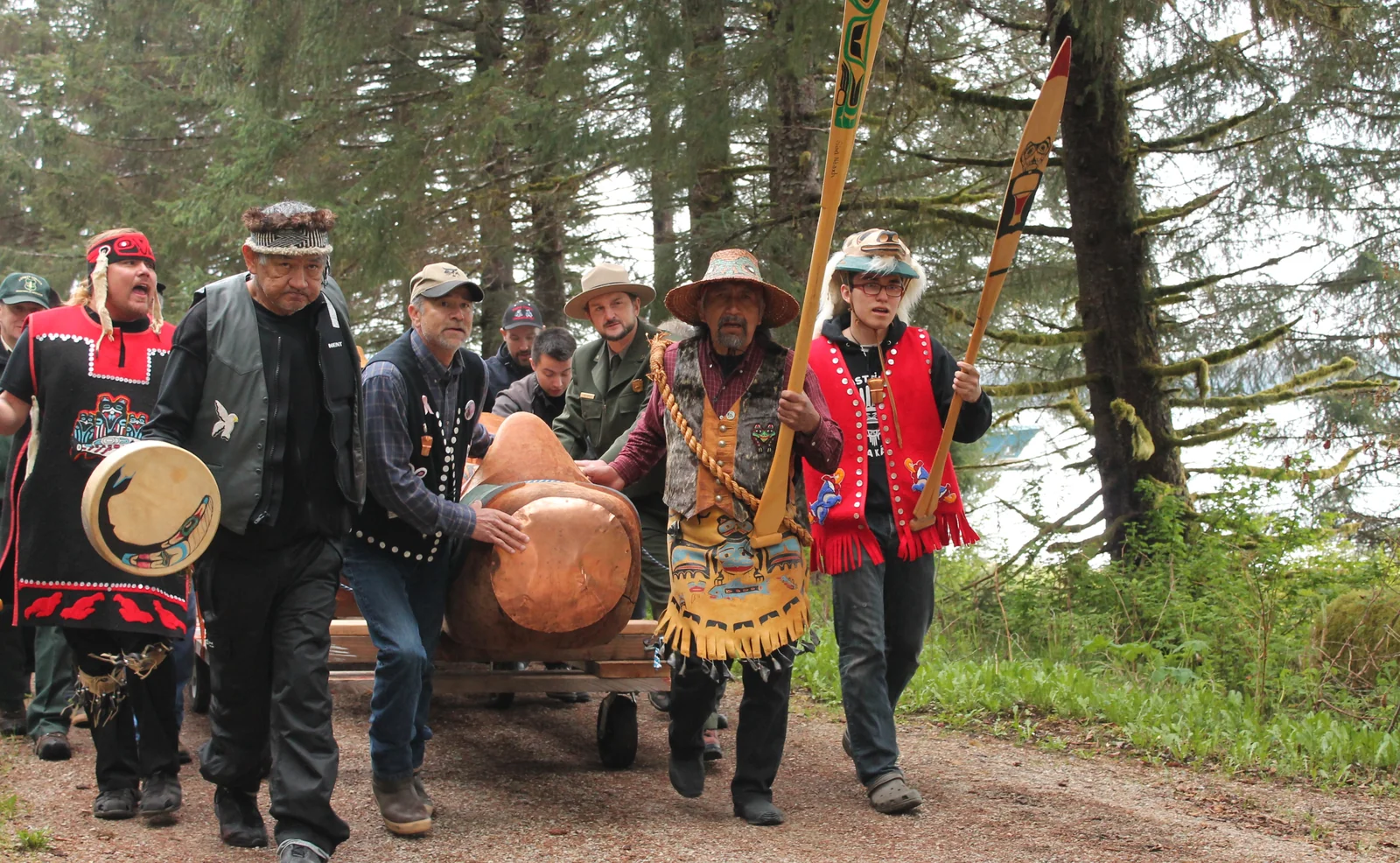 A group of Huna Tlingit men in ceremonial regalia carry a totem pole along a forest path