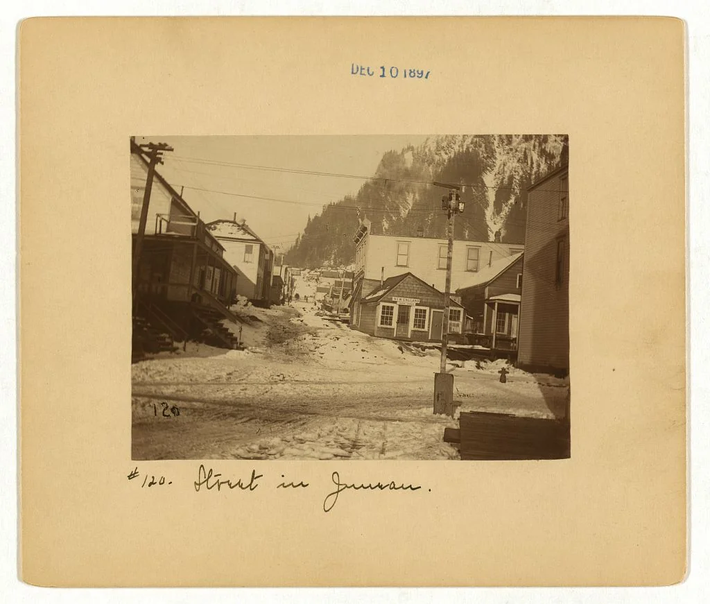 1897 sepia photograph of a muddy wooden-storefront street in early Juneau, Alaska