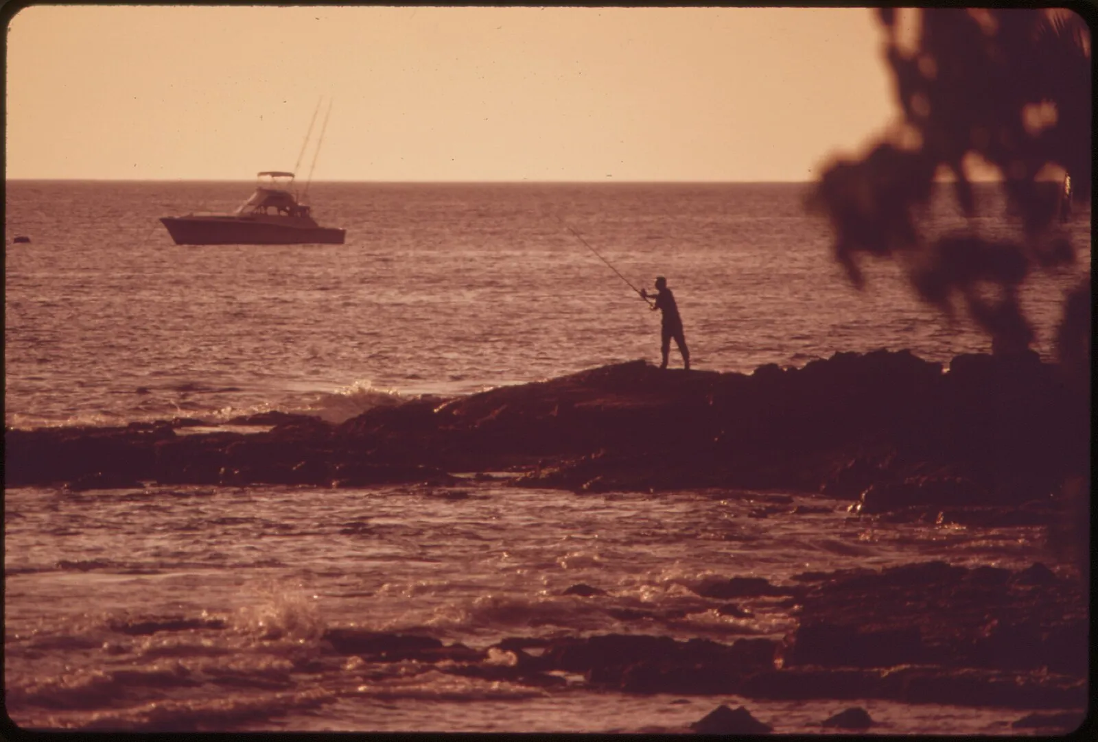 Vintage silhouette of fisherman on lava rocks at sunset with charter boat off Kona coast