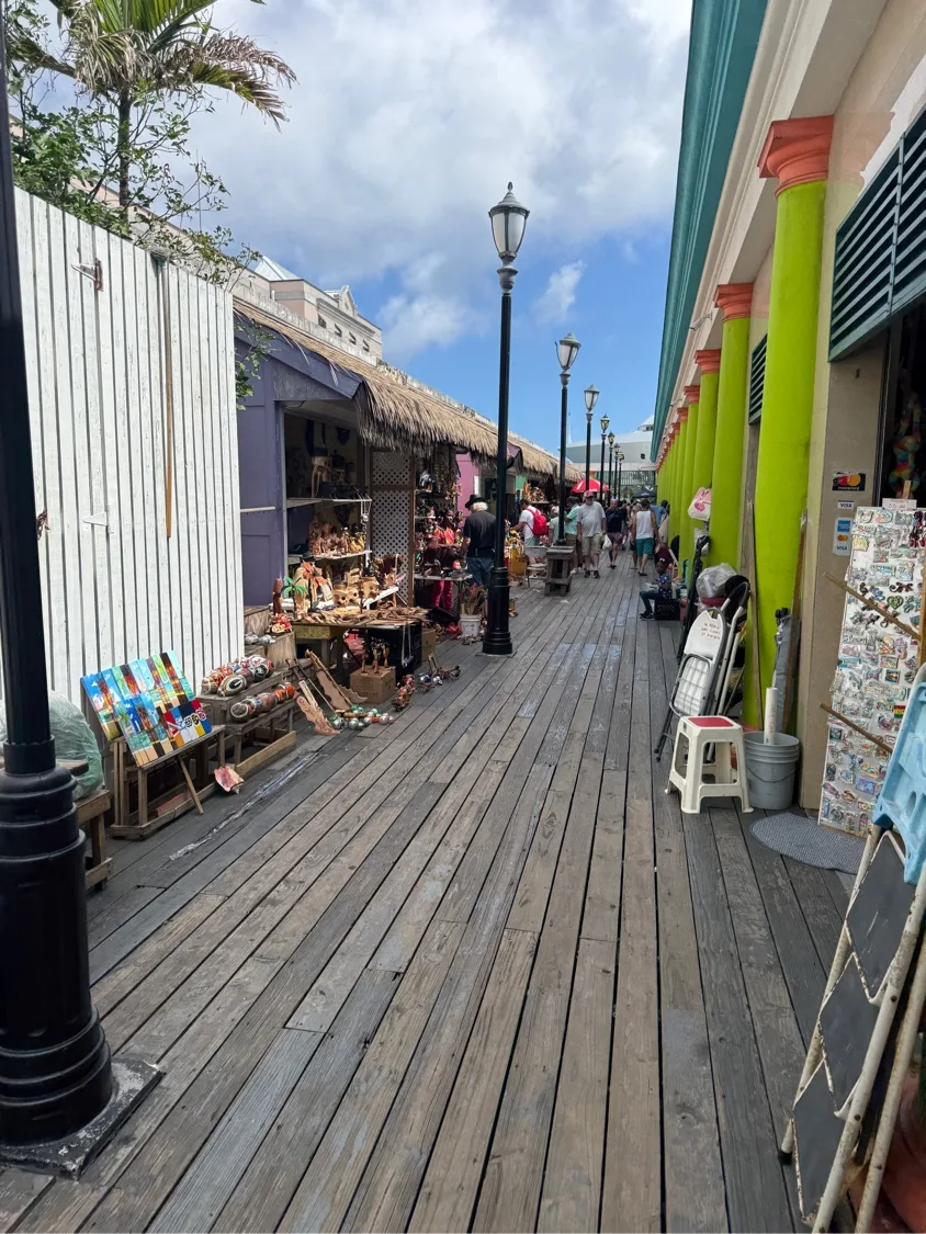 Inside the Straw Market — a wooden-board promenade lined with thatched-roof vendor stalls displaying wood carvings, dolls, and Bahamian crafts under iron lampposts between flamingo-pink and mint-green buildings