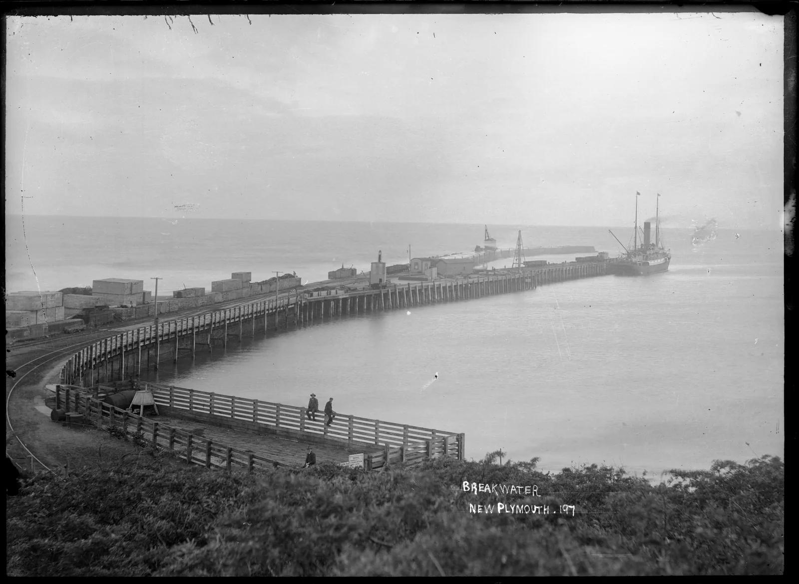 Black and white photograph from about 1910 showing the timber breakwater wharf at New Plymouth curving out from the shore, a railway line and goods wagons running along it, and a steamer berthed at the far end with smoke rising from its funnel