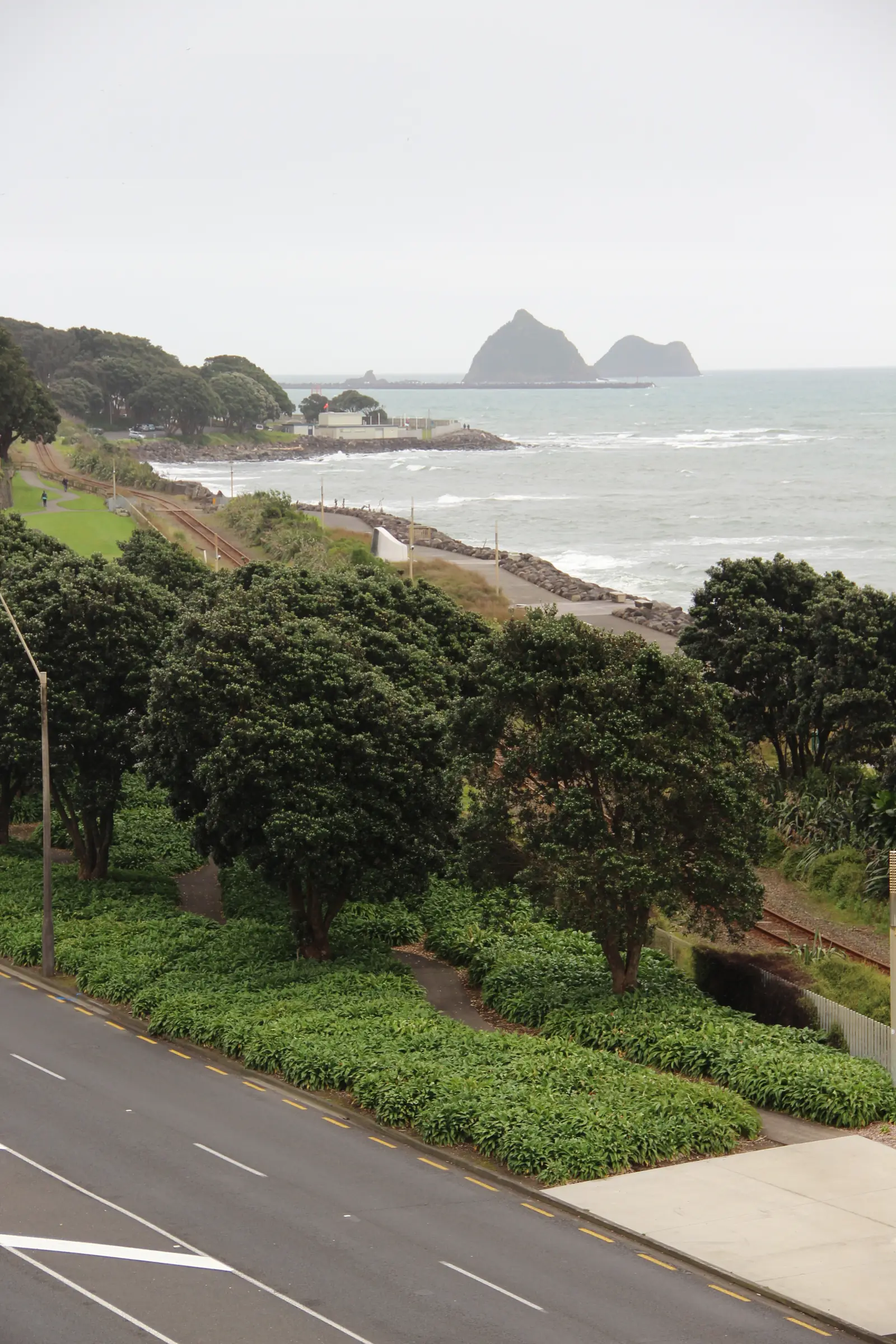 A section of the New Plymouth Coastal Walkway seen from above a coastal road, curving along a rocky shore past pohutukawa trees and a railway line, with the port breakwater and the Sugar Loaf Islands on the horizon