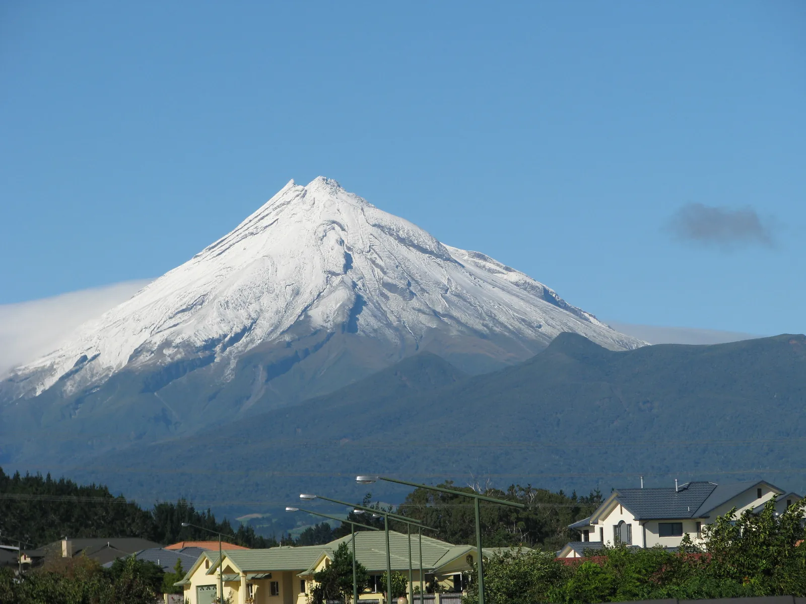 The snow-streaked cone of Taranaki Maunga rising above the rooftops, power lines and street lamps of suburban New Plymouth under a clear blue sky, with a low bank of cloud clinging to the mountain's lower left flank