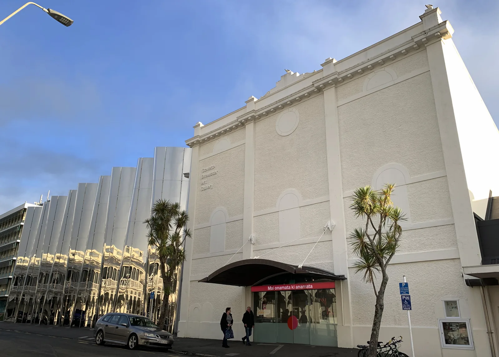 The Govett-Brewster Art Gallery and Len Lye Centre on Queen Street, New Plymouth, its curved mirrored stainless-steel columns reflecting the heritage buildings opposite alongside the gallery's plain cream facade