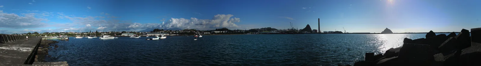 Wide panorama across the sheltered water inside Port Taranaki, with yachts and launches at moorings, the town rising behind, the power station chimney and Paritūtū on the skyline, and a rock breakwater in the right foreground