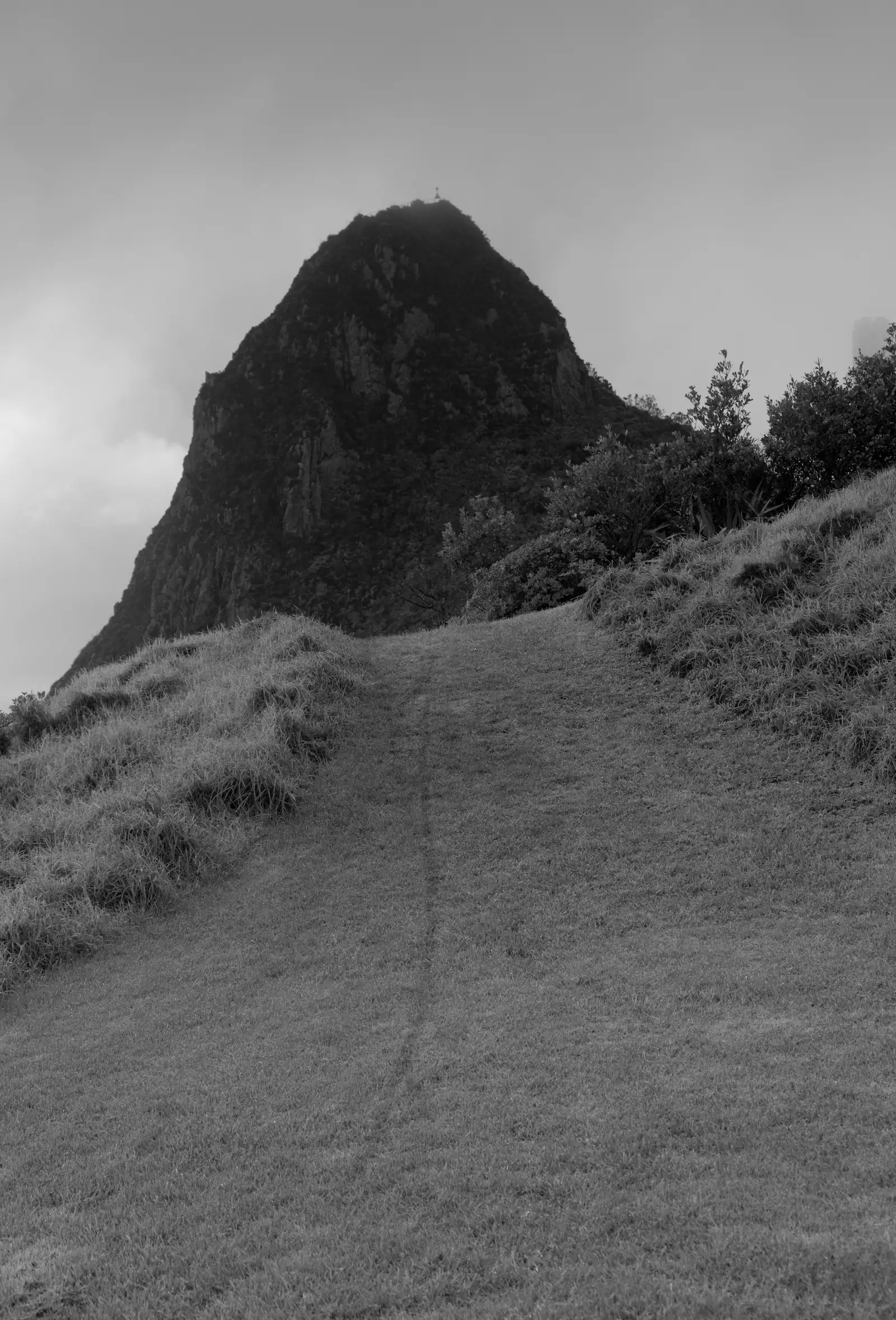 Monochrome photograph of Paritūtū, a steep conical volcanic remnant rising above a grassed slope, with a single figure just visible standing on the summit