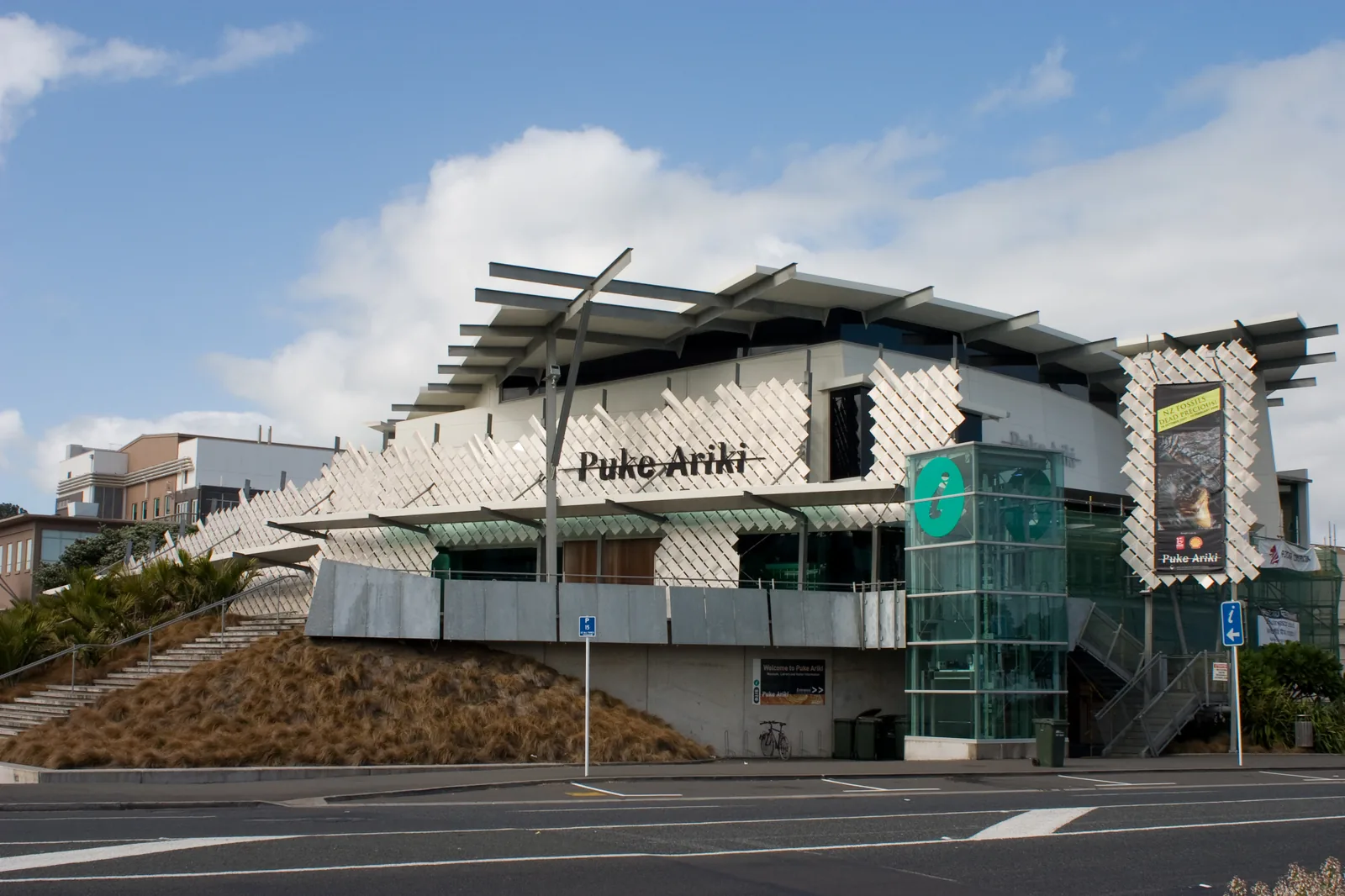 The angular white exterior of Puke Ariki museum and library in New Plymouth, with its name in large lettering across a lattice-clad facade, a glass lift tower, and steps leading up from the street