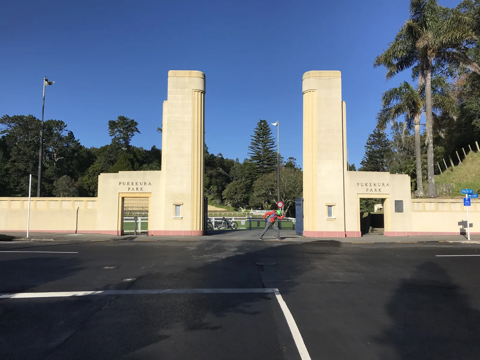 The Art Deco entrance pylons of Pukekura Park, New Plymouth, two tall cream fluted towers flanking pedestrian archways lettered PUKEKURA PARK, with native bush and a Norfolk pine behind