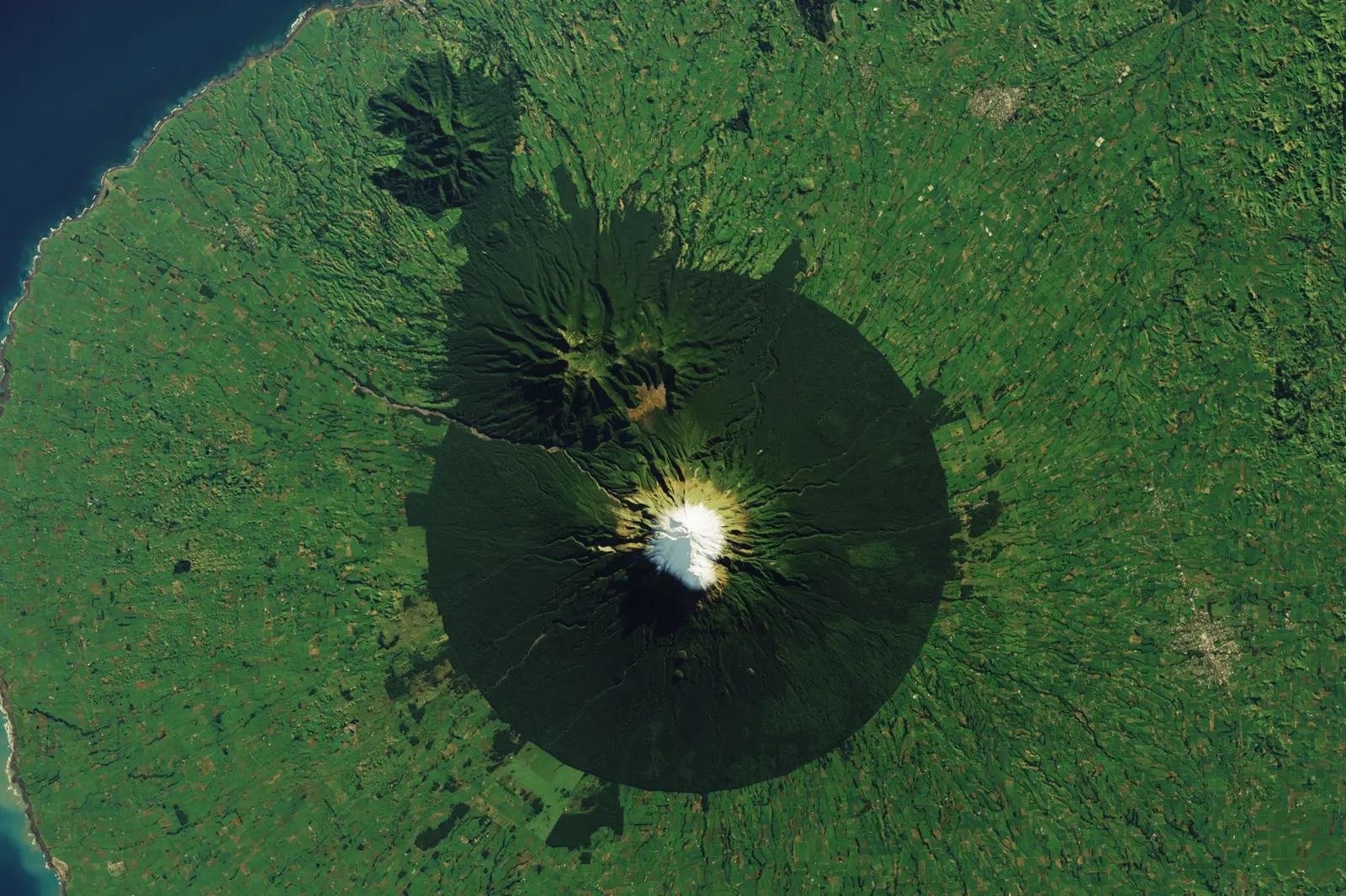 Landsat satellite image of Taranaki Maunga showing a near-perfect dark circle of protected forest around the snow-capped cone, with bright green dairy pasture pressed right up to the boundary and the Tasman coastline at the upper left