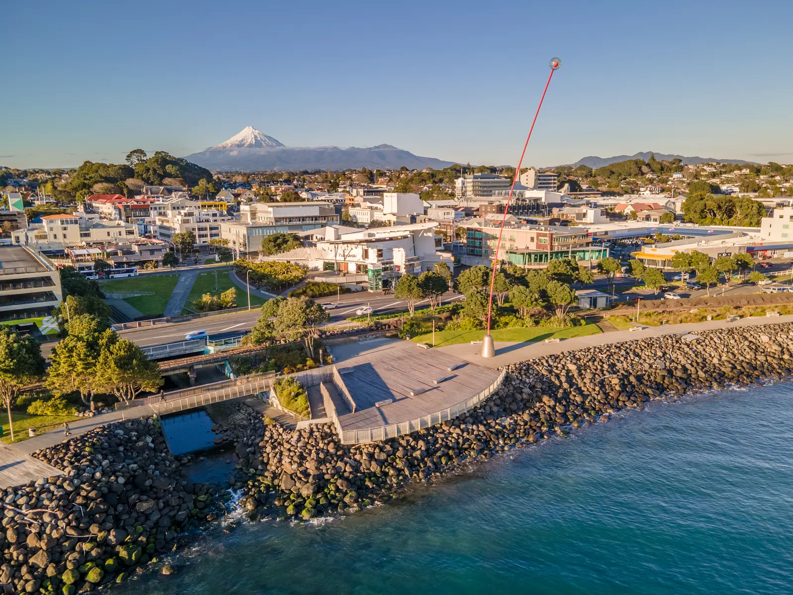 Aerial view of the New Plymouth waterfront at golden hour showing the red Wind Wand sculpture, the curved timber viewing platform, the rocky foreshore, the low city centre and the snow-capped cone of Taranaki Maunga rising behind