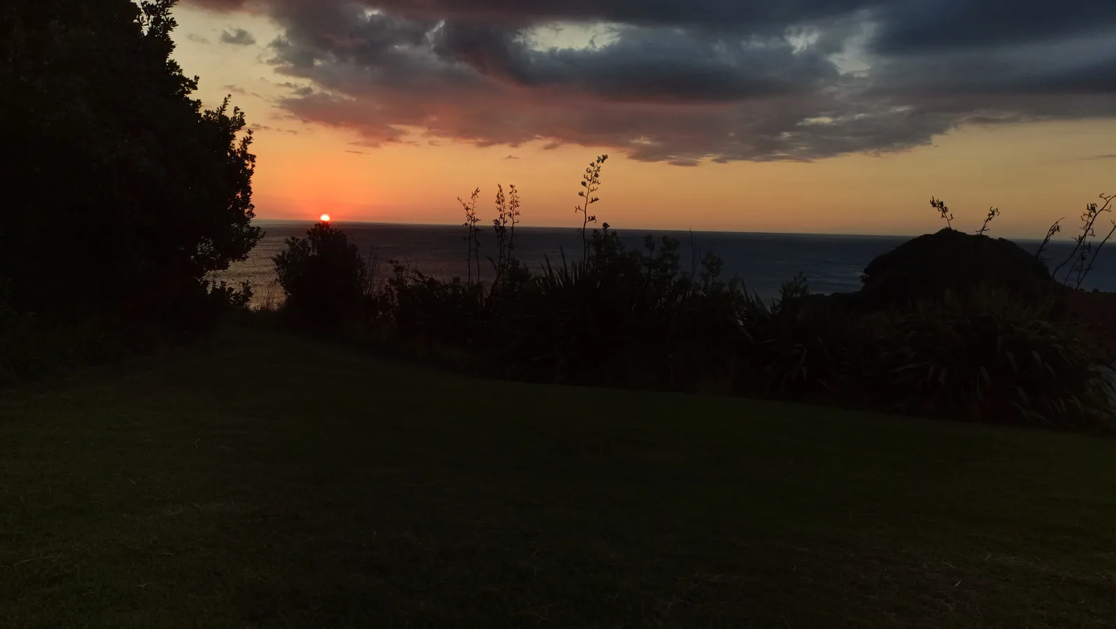 A vivid orange and pink sunset over the sea at New Plymouth, with the dark silhouette of the coastline and offshore rocks in the foreground