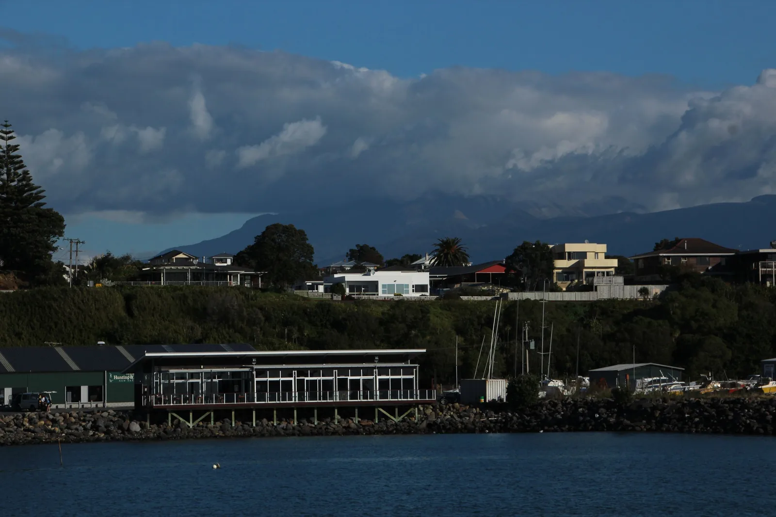 The New Plymouth waterfront in bright sun beneath a heavy bank of cloud that completely conceals Taranaki Maunga, with only the dark lower ridges of the mountain visible behind the houses and the marina buildings