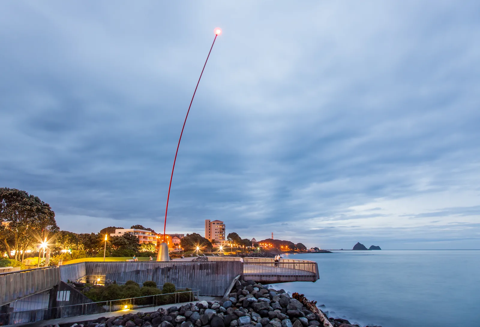 The Wind Wand at dusk on the New Plymouth foreshore, a slender red mast curving against a blue-grey sky with its lit red sphere at the tip, the town lights along the shore and the Sugar Loaf Islands on the horizon