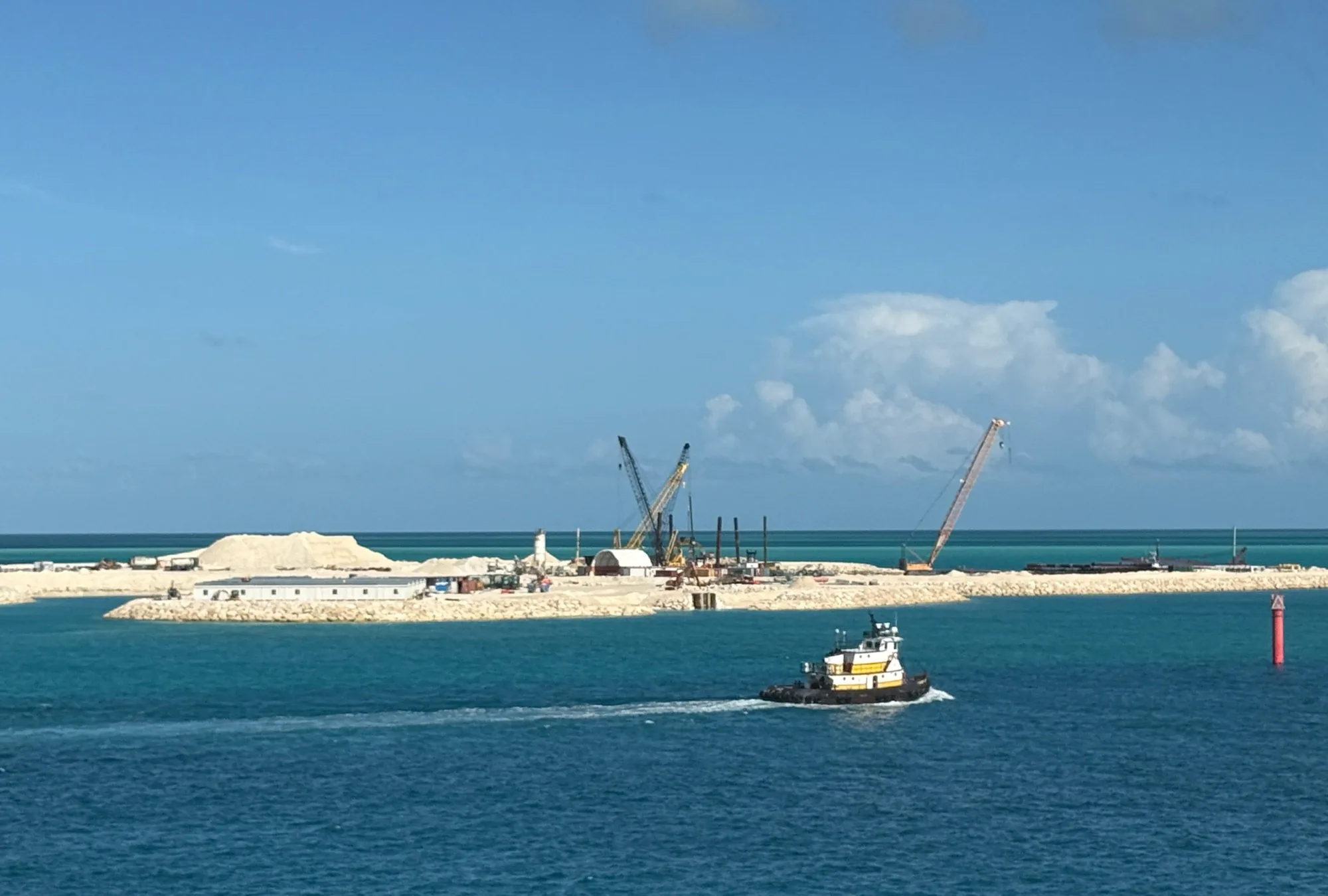 Active construction at Ocean Cay's south end: two lattice-boom cranes on a new rock-armoured spit, harbor tugboat crossing the channel