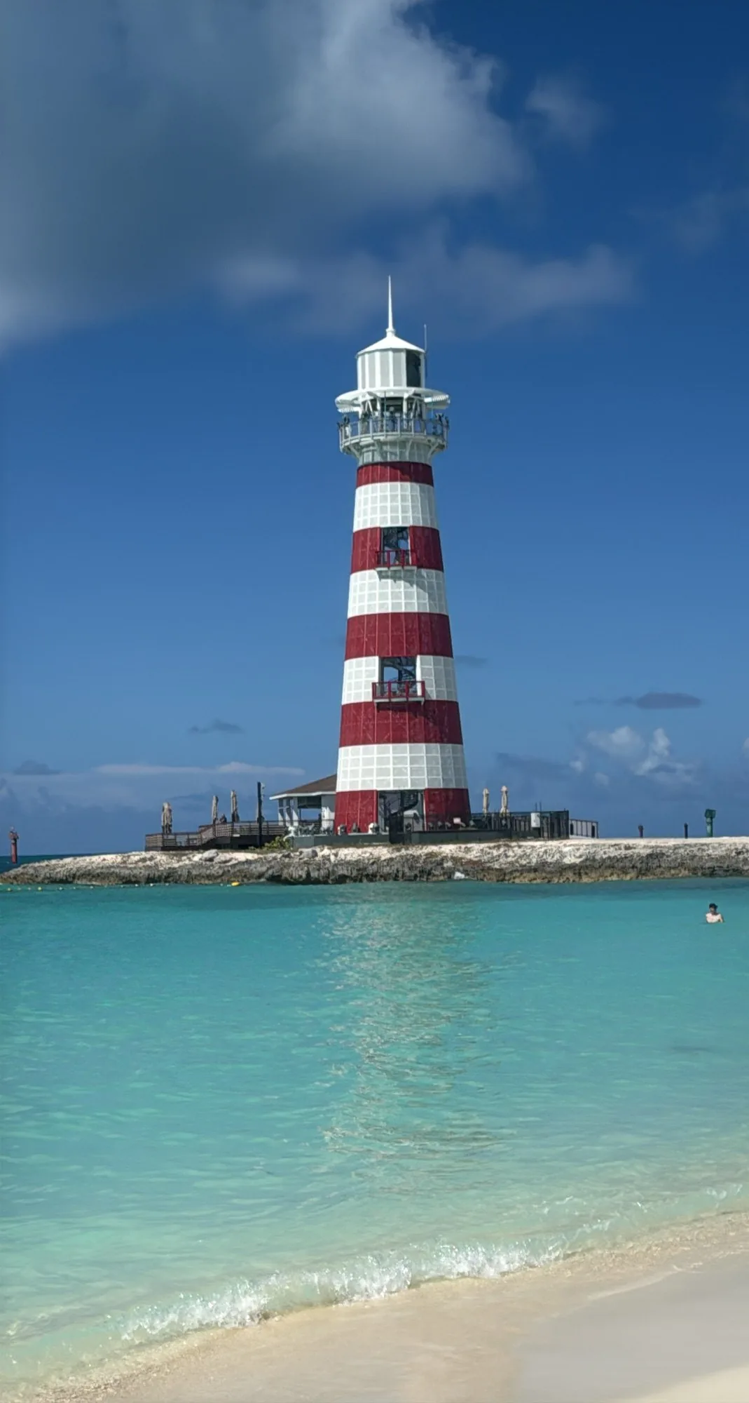 Ocean Cay lighthouse on its rocky spit seen from the swim beach across glassy turquoise shallows, single swimmer in the water